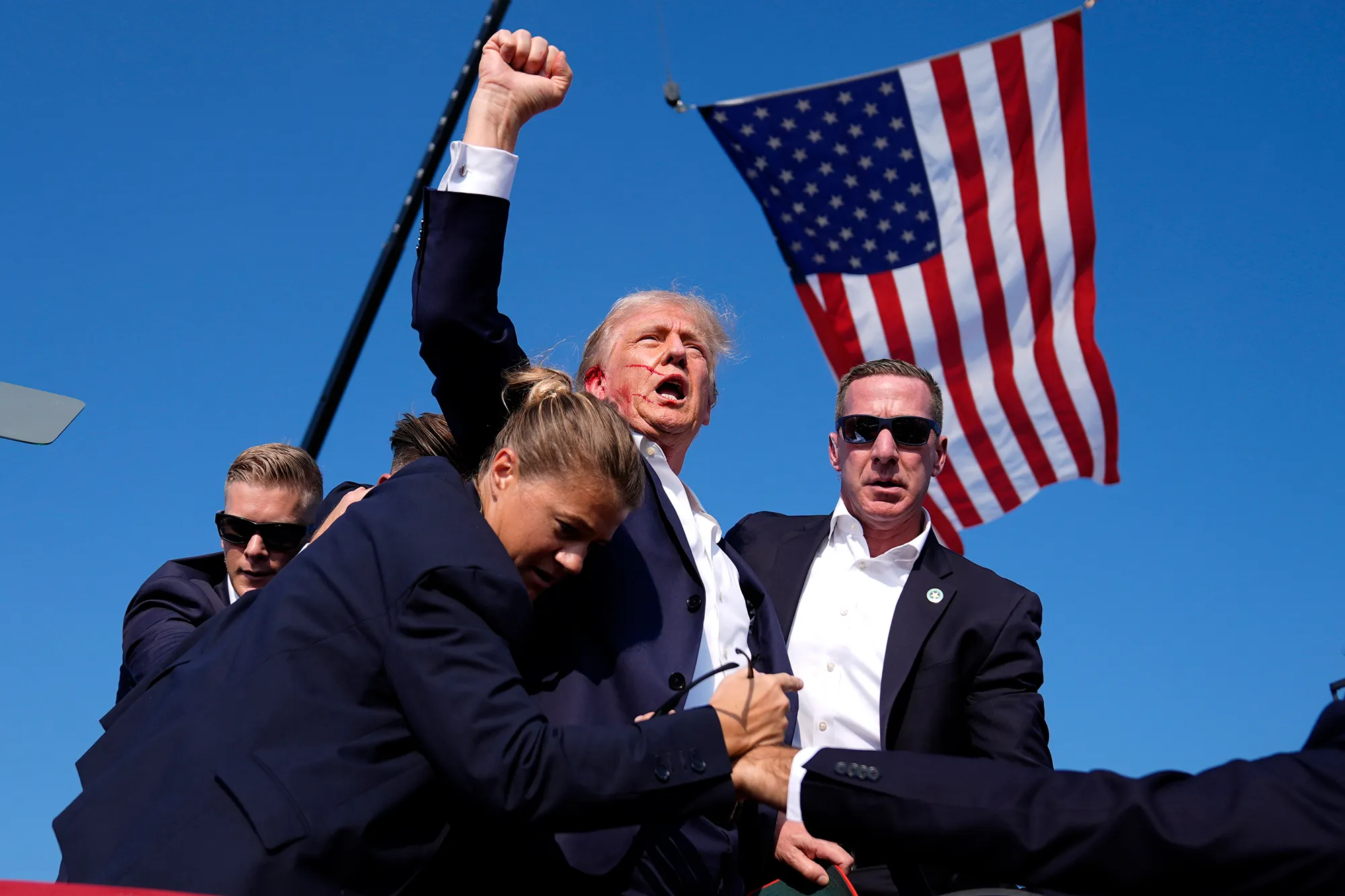 Donald Trump with a bloody ear, raising fist in the air, surrounded by secret service